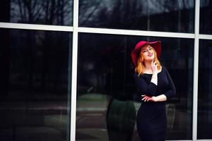 Porträt eines modischen, rothaarigen Mädchens mit rotem Hut und schwarzem Kleid mit hellem Make-up, das gegen ein großes Fenster gestellt wurde. Foto getönte Instagram-Filter.