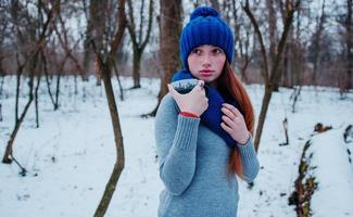 Porträt eines jungen Mädchens mit roten Haaren und Sommersprossen, das am Wintertag eine blau gestrickte Wollmütze und einen Schal mit einer Tasse Tee trägt. foto