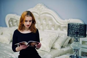 rothaariges Mädchen auf schwarzem Kleid, das auf dem Bett sitzt und Modezeitschriften liest. Foto getönte Instagram-Filter.