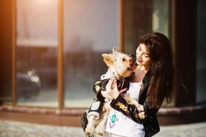 Brünette Zigeunerin mit Yorkshire-Terrier-Hund posierte gegen Haus mit großen Fenstern. Model Wear auf Lederjacke und T-Shirt mit Ornament, Hose. foto