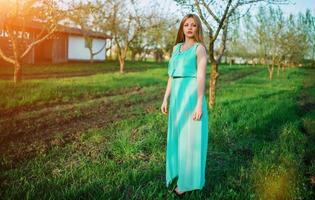 Frau in einem wunderschönen langen türkisfarbenen Kleid posiert auf einer Wiese im Obstgarten. foto