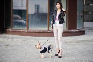 Brünette Zigeunerin mit Yorkshire-Terrier-Hund posierte gegen Haus mit großen Fenstern. modelwear auf lederjacke und t-shirt mit verzierung, hose und schuhe mit high heels. foto