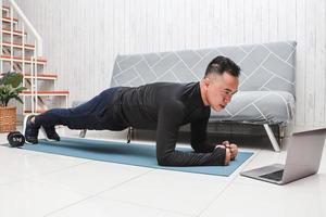 asiatischer mann zu hause, der übungen macht und sich aufwärmt, während er sich eine online-video-fitnessstunde auf dem laptop ansieht foto