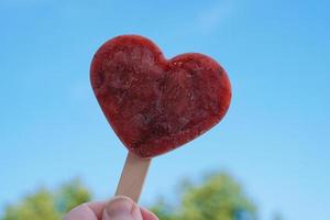 Hand, die herzförmiges Eis am Stiel oder Eis am Stiel gegen den klaren blauen Himmel hält foto