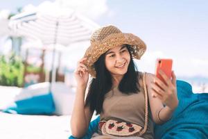reise asiatin entspannen sich im strandcafé und nutzen das handy für videoanrufe über das online-internet. foto