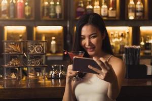 asiatische Frau, die ein Foto von sich macht, während sie Whiskey an der Bar trinkt.
