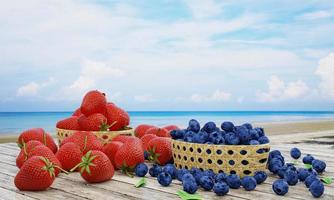 frische Erdbeeren und Heidelbeeren in einem Bambuskorb. Beere auf einer Holzoberfläche. Hintergrundlandschaft ist Meer und Strand, blauer Himmel und weiße Wolken, Landschaft und Sonnenlicht am Morgen. 3D-Rendering. foto