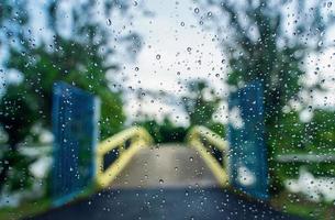 Regentropfen auf der Oberfläche von Autoglas mit unscharfer grüner Natur und Brückenhintergrund durch Fensterglas des Autos, das von Regentropfen bedeckt ist. Frische nach Regen. Nasse Windschutzscheibe aus dem Auto heraus geschossen. foto