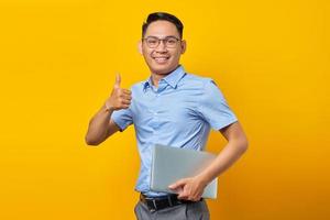 Porträt eines lächelnden, gutaussehenden asiatischen Mannes mit Brille, der einen Laptop hält und eine Geste mit dem Daumen nach oben macht, genehmigen Sie die gute Sache, die auf gelbem Hintergrund isoliert ist. geschäftsmann- und unternehmerkonzept foto