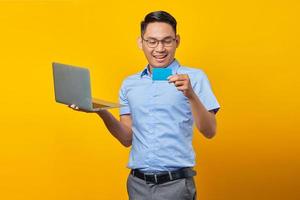 Aufgeregter junger asiatischer Mann mit Brille, der einen Laptop hält und eine Kreditkarte isoliert auf gelbem Hintergrund sieht. geschäftsmann- und unternehmerkonzept foto