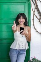glückliche hispanische frau mit dem smartphone, das daumen nach oben gestikuliert foto