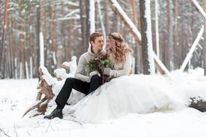 Süßes Paar, das sich in einen Blumenstrauß verliebt, sitzt auf dem Baumstamm im Hintergrund des Winterwaldes. Kunstwerk. Winterhochzeit. Platz kopieren foto