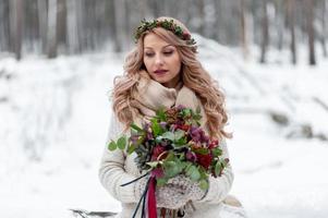 ein junges Mädchen slawischen Aussehens mit einem Kranz aus Wildblumen. Schöne blonde Braut hält einen Blumenstrauß im Winterhintergrund. foto