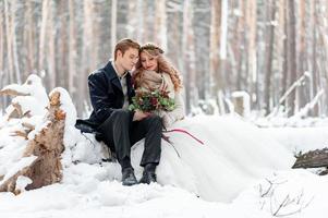 Süßes Paar, das sich in einen Blumenstrauß verliebt, sitzt auf dem Baumstamm im Hintergrund des Winterwaldes. Kunstwerk. Winterhochzeit. Platz kopieren foto