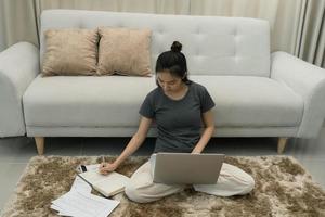 asiatische frau sitzt auf einem teppich im wohnzimmer und arbeitet an einem computer, während sie notizen in ein notizbuch schreibt. foto