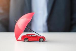 geschäftsmannhand, die regenschirm hält und rotes autospielzeug auf dem tisch abdeckt. autoversicherung, garantie, reparatur, finanz-, bank- und geldkonzept foto