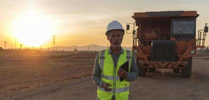 leitender ingenieur, der eine sicherheitsweste und einen helm trägt und ein digitales tablet auf der baustelle hält, ein älterer arbeiter, der arbeitet, hat einen großen lastwagen als hintergrund foto