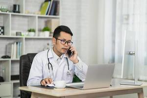 asiatischer arzt, der mobiltelefon anruft und den patienten per videoanruf im heimbüro mit laptop konsultiert. telemedizin- und gesundheitskonzept foto