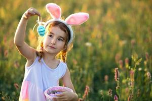 süßes lustiges Mädchen mit bemalten Ostereiern im Frühling in der Natur auf einem Feld mit goldenem Sonnenlicht und Blumen. osterferien, osterhase mit ohren, bunte eier in einem korb. Lebensstil foto
