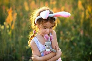 süßes 5-jähriges Mädchen mit Hasenohren umarmt sanft ein Spielzeugkaninchen in der Natur auf einem blühenden Feld im Sommer mit goldenem Sonnenlicht. ostern, osterhase, kindheit, glückliches kind, frühling. foto