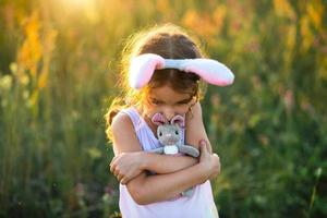 süßes 5-jähriges Mädchen mit Hasenohren umarmt sanft ein Spielzeugkaninchen in der Natur auf einem blühenden Feld im Sommer mit goldenem Sonnenlicht. ostern, osterhase, kindheit, glückliches kind, frühling. foto