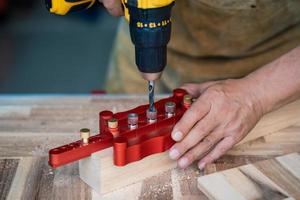 Der Zimmermann verwendet einen Bohrer und eine Zentrierdübellehre oder ein Taschenlochlehrenwerkzeug, um starke Verbindungen auf Holzplatten herzustellen. Holzbearbeitungskonzept. Selektiver Fokus. foto