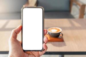 Hand, die Smartphone-Gerät mit leerem Bildschirm im Café hält, unscharfer Kaffeetassenhintergrund, Mock-up-Konzept foto