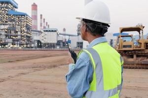 ein leitender ingenieur mit helm und sicherheitsweste mit tragbarem walkie-talkie ist im hintergrund des kraftwerks zu sehen. foto