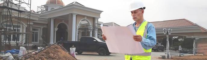 Bauingenieur mittleren Alters mit Schutzhelm mit Projekt in der Hand. asiatische Älteste, die auf der Baustelle stehen foto