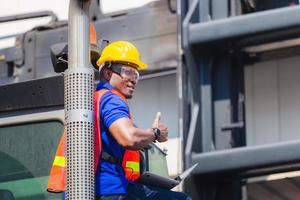 arbeiter in helm und sicherheitsweste, der einen laptop hält, der auf containerstaplern steht, steuert das laden von containern aus der fracht, lächelnder mann und gibt daumen hoch foto