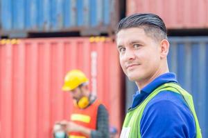 Vorarbeiter mit Blick auf die Kamera in Industriecontainern mit verschwommenem Ingenieurmann, der im Hintergrund ein Mobiltelefon benutzt foto