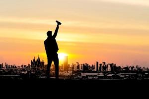 Silhouette eines erfolgreichen Geschäftsmannes, der die Trophäe mit dem Sonnenuntergangshintergrund des Stadtabendhimmels aufhebt und den Mann feiert, der die Siegerpokaltrophäe hält foto