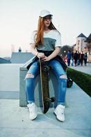 Junges urbanes Mädchen im Teenageralter mit Skateboard, Brille, Mütze und zerrissenen Jeans am Abend im Skatepark. foto