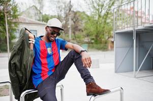 stylische afroamerikanische jungenbekleidung an kappe, fußball-t-shirt und sonnenbrille posiert auf stahlgeländern. schwarzes Sportlerporträt. foto