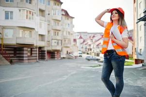 ingenieurbaumeisterin in uniformweste und orangefarbenem schutzhelm halten geschäftszeichnungspapierrolle gegen neues gebäude. Immobilien-Wohnblock-Thema. foto