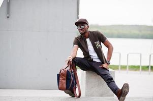 porträt des sitzenden stilvollen afroamerikaners, der auf sonnenbrille und kappe im freien trägt. Straßenmode schwarzer Mann. foto