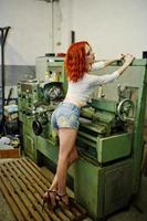 rothaarige mädchen tragen kurze jeansshorts und weiße bluse, die an der industriemaschine in der fabrik gestellt wurden. foto