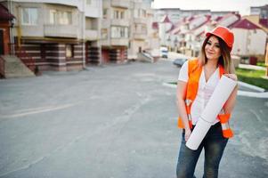 ingenieurbaumeisterin in uniformweste und orangefarbenem schutzhelm halten geschäftszeichnungspapierrolle gegen neues gebäude. Immobilien-Wohnblock-Thema. foto