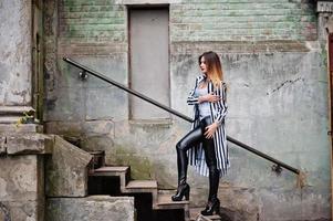 Modischer Frauenlook mit schwarz-weiß gestreifter Anzugjacke, Lederhose, posiert auf der alten Straße auf Eisentreppen. Konzept des Modemädchens. foto