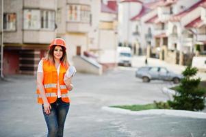 ingenieurbaumeisterin in uniformweste und orangefarbenem schutzhelm halten geschäftszeichnungspapierrolle gegen neues gebäude. Immobilien-Wohnblock-Thema. foto