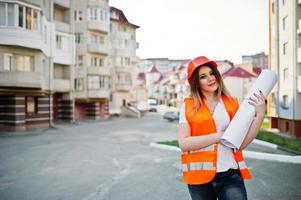 ingenieurbaumeisterin in uniformweste und orangefarbenem schutzhelm halten geschäftszeichnungspapierrolle gegen neues gebäude. Immobilien-Wohnblock-Thema. foto