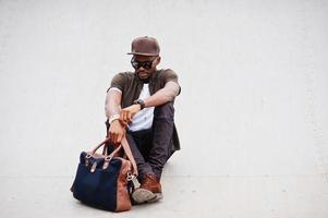 porträt des sitzenden stilvollen afroamerikaners, der auf sonnenbrille und kappe mit handtasche im freien trägt. Straßenmode schwarzer Mann. foto