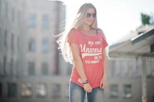 stilvolle, glückliche blonde frau trägt jeans, sonnenbrille und t-shirt, das bei sonnigem wetter auf der straße posiert. Mode urbanes Modellporträt. foto