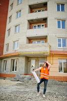 ingenieurbaumeisterin in uniformweste und orangefarbenem schutzhelm halten geschäftszeichnungspapierrolle gegen neues gebäude. Immobilien-Wohnblock-Thema. foto