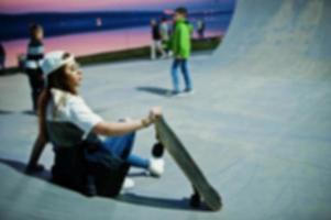 Verschwommenes Foto eines jungen urbanen Mädchens im Teenageralter mit Skateboard, Brille, Mütze und zerrissenen Jeans im Skatepark am Abend.