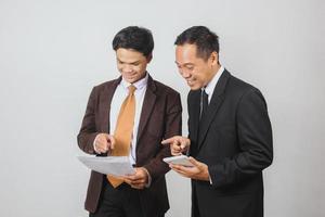 Zwei asiatische Geschäftsleute im Anzug lächeln mit einem Smartphone, während sie auf einen Papierbericht zeigen foto