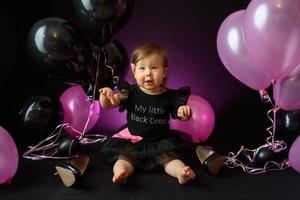 Geburtstagsfeiertag des ersten Jahres des Babymädchens. Ballons und Urlaub drinnen. Geburtstag des Kindes. kleines hübsches Mädchen in ihrem ersten schwarzen Kleid foto