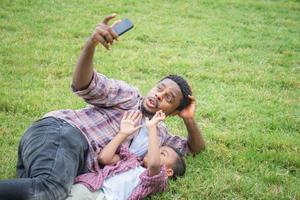 glücklicher vater mit kleinem sohn, der selfie-foto mit smartphone macht, fröhlicher afroamerikanischer vater und söhne, die im park spielen foto