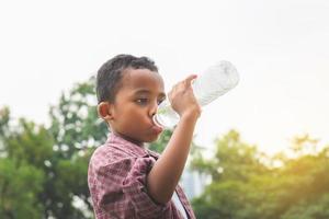 Fröhlicher afrikanisch-amerikanischer Junge trinkt Wasser, nachdem er im Park gespielt hat foto