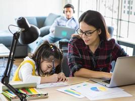 muti ethnische familie, vater und mutter und junge kleine tochter bleiben zusammen im wohnzimmer. mutter arbeitet von zu hause aus mit laptop-notebook-computer, während sie mädchen beibringt, hausaufgaben zu machen. neues normales Konzept foto